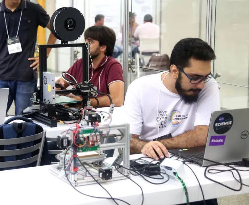 Bancada de prototipagem da INOVAeye, com impressora 3D e placas eletrônicas em evento de tecnologia